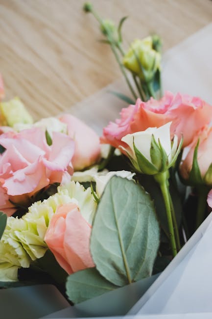 A close-up view of a bouquet featuring pale pink and cream lisianthus flowers with delicate, ruffled petals, accompanied by fresh green leaves. The arrangement is partially wrapped in elegant, semi-tr