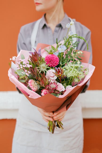 A person dressed in a light grey shirt and white apron, holding a large floral bouquet wrapped in soft pink and brown paper. The bouquet features a variety of fresh flowers, including vibrant pink ger