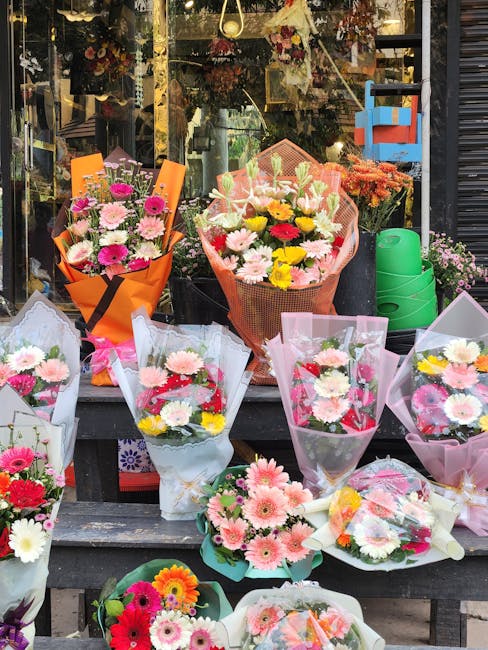 A variety of fresh flower bouquets displayed outside a shop window, featuring an assortment of colorful blooms including pink daisies, yellow chrysanthemums, white lilies, and purple asters. The bouqu
