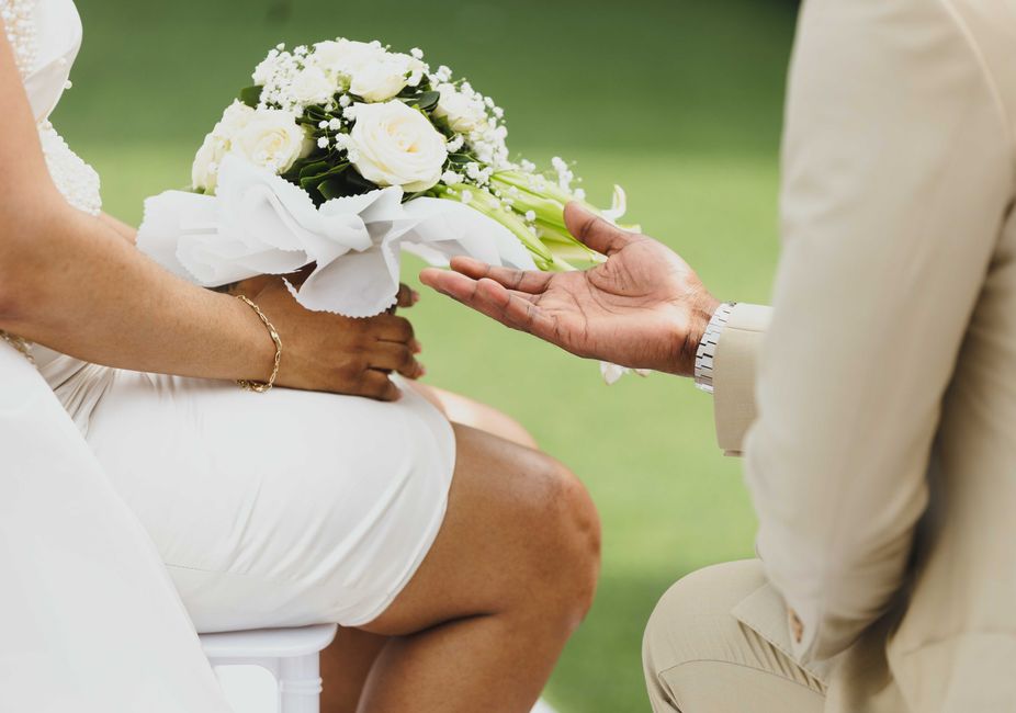 A close-up image of a wedding bouquet featuring white roses, baby's breath, and greenery, held by a bride dressed in white with a gold bracelet. The bouquet is wrapped in white tissue paper and is bei
