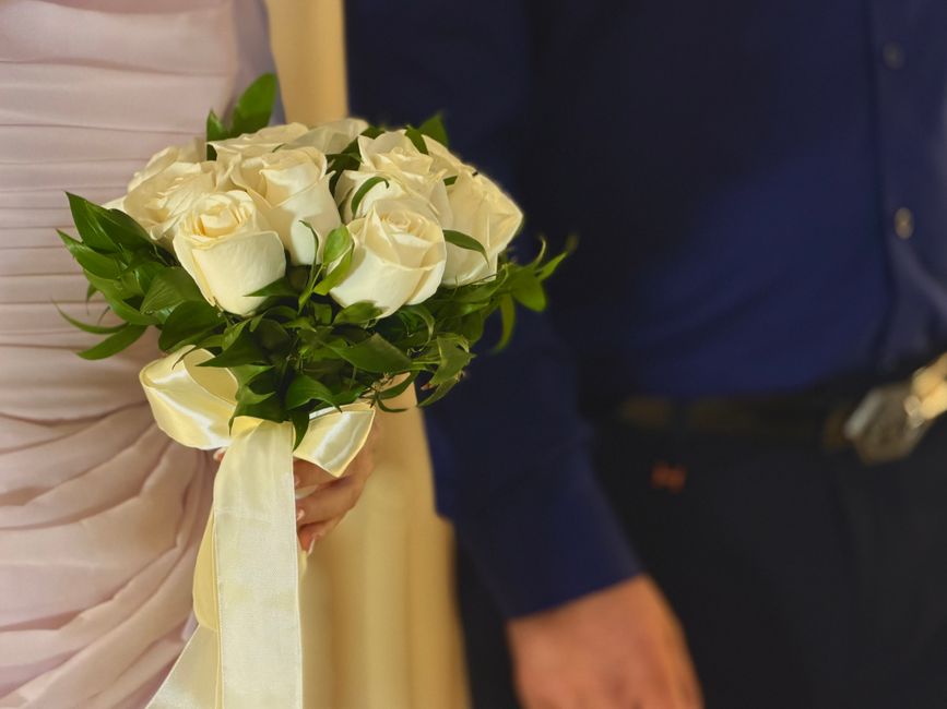 A close-up of a bridal bouquet held by a bride, featuring a cluster of creamy white roses with lush green foliage. The roses have smooth, velvety petals arranged tightly, creating a rounded shape. The