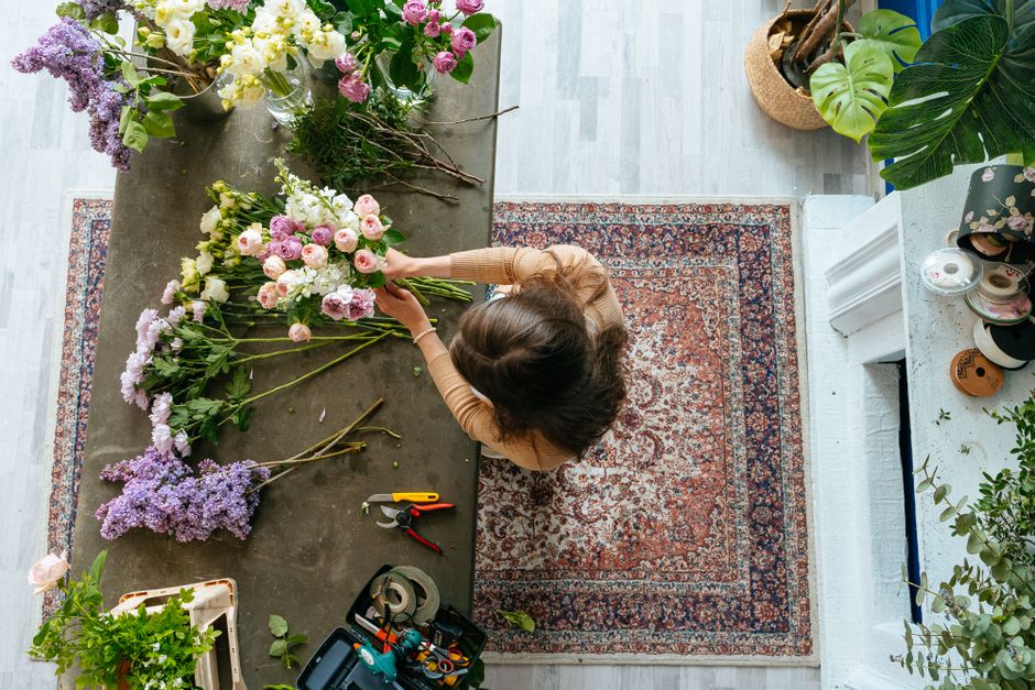 A professional florist working on floral arrangements at a grey worktable, surrounded by various fresh flowers including pink roses, purple lilacs, and white blooms. The florist is placing the flowers