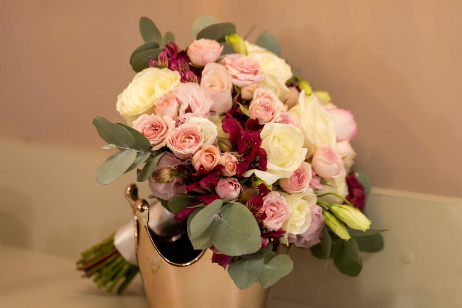 A bouquet of fresh flowers arranged in a rounded, lush style, featuring white roses, pale pink spray roses, and deep burgundy blooms, accented with eucalyptus leaves. The bouquet is wrapped with beige