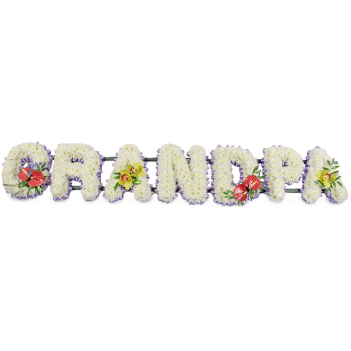 This image shows a striking, custom funeral tribute that clearly spells the word "GRANDPA" in substantial, block-style letters arranged in a straight horizontal line, ideal for placing along the front of a coffin or on stands in a Hither Green funeral chapel. Each letter is completely covered with tight clusters of small white chrysanthemums, their petals forming a soft, domed surface that suggests peace, purity, and gentle remembrance. The flowers are packed so closely that no gaps are visible, creating a neat, professional finish that reflects the experienced hand of a local florist used to preparing family tributes for services around Hither Green, Lee, and Blackheath. A subtle edging of pale lavender ribbon lines the bottom of every letter, just visible beneath the white blooms like a delicate halo, adding a pastel contrast without disturbing the calm mood. Discreet dark green bars link the letters together in one stable unit, hidden from the front but providing vital support. At four points along the tribute, vibrant accent sprays add colour and personality: the "G" and "D" are adorned with bold red anthuriums, their glossy, heart-shaped petals and protruding yellow spadices standing out vividly against the white, surrounded by polished greenery and hints of tiny purple flowers. The first and the final "A" each carry a cluster of yellow-green orchids, their sculpted petals and subtle markings conveying elegance and enduring love. The overall composition feels both formal and deeply personal, a loving tribute that speaks of respect, gratitude, and cherished memories of a grandfather's presence across years of family life.